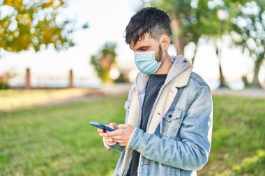 Parkta akıllı telefon kullanan tıbbi maske takan genç bir İspanyol.