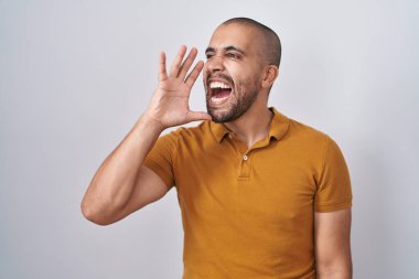 Hispanic man with beard standing over white background shouting and screaming loud to side with hand on mouth. communication concept. 