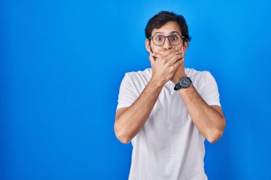 Mavi arka planda duran yakışıklı latin adam yanlışlıkla elleri ile ağzını kapatırken şok oldu. gizli kavram. 