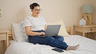 Young beautiful hispanic woman using laptop sitting on bed at bedroom
