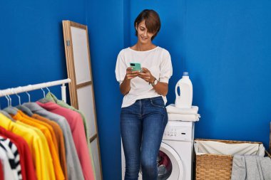 Çamaşır odasında çamaşır makinesini bekleyen akıllı telefon kullanan genç ve güzel bir İspanyol kadın.