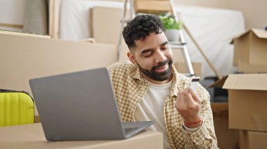 Genç İspanyol adam dizüstü bilgisayarı ve kredi kartıyla alışveriş yapıyor. Yeni evinde yerde oturuyor.