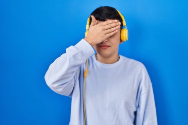 İkili olmayan kişi müzik dinlerken kulaklık takıyor, gözleri elle örtüyor, ciddi ve üzgün görünüyor. Görme, saklama ve reddetme konsepti 