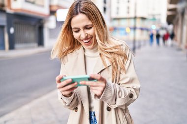 Genç sarışın kadın gülümsüyor. Sokakta akıllı telefondan videolar izliyor.