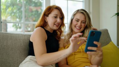 Two women using smartphone sitting on sofa at home