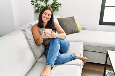 Genç ve güzel İspanyol kadın evdeki kanepede oturup kahve içiyor.