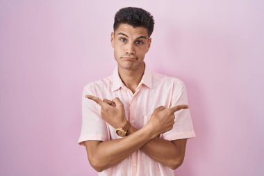 Young hispanic man standing over pink background pointing to both sides with fingers, different direction disagree 