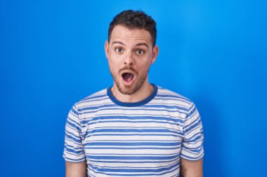 Young hispanic man standing over blue background afraid and shocked with surprise and amazed expression, fear and excited face. 