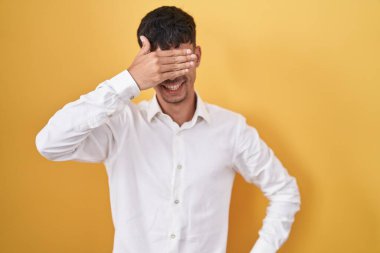 Young hispanic man standing over yellow background smiling and laughing with hand on face covering eyes for surprise. blind concept. 