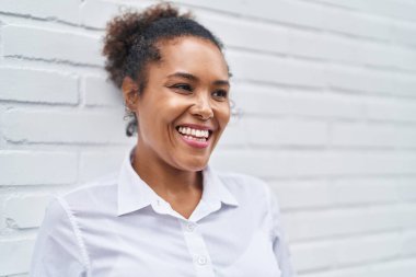 Afro-Amerikalı kadın gülerek beyaz tuğla arka planına bakıyor.