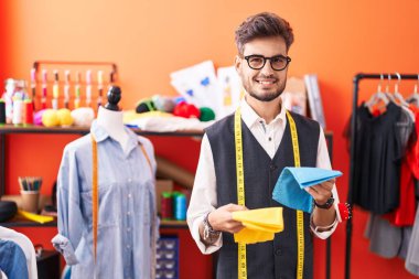Young hispanic man tailor smiling confident holding cloths at atelier
