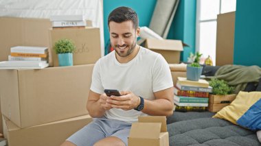 Genç İspanyol adam akıllı telefon kullanıyor. Yeni evindeki kanepede oturuyor.