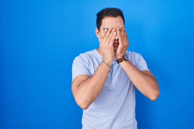 Mavi arka planda duran İspanyol genç adam yorgunluk ve baş ağrısı için gözlerini ovuşturuyor, uykulu ve yorgun ifade. görme sorunu 