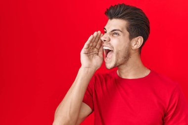 Young hispanic man standing over red background shouting and screaming loud to side with hand on mouth. communication concept. 