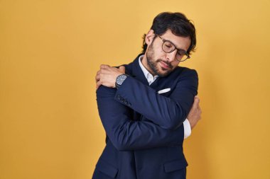 Sarı arka planda duran yakışıklı Latin adam kendine sarılıyor mutlu ve pozitif, kendinden emin gülümsüyor. Sevgi ve özgüven. 