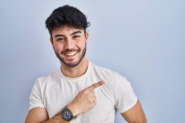 Hispanic man with beard standing over white background cheerful with a smile on face pointing with hand and finger up to the side with happy and natural expression 