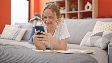 Evdeki kanepede uzanmış akıllı telefon kullanan genç sarışın kadın.