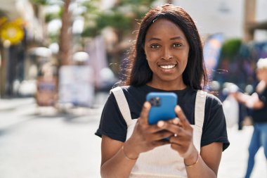 Genç Afro-Amerikalı kadın sokakta akıllı telefon kullanarak gülümsüyor.