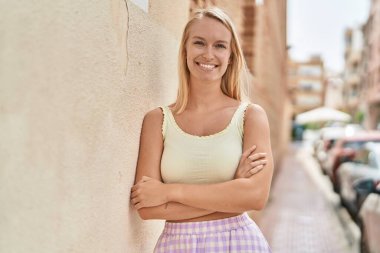 Genç sarışın kadın gülümsüyor. Kollarını kavuşturup caddede duruyor.