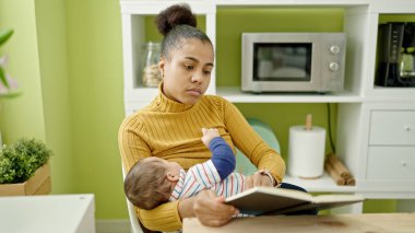 Anne ve oğul yemek odasında bebek emzirirken kitap okuyorlar.