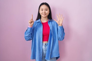 Young asian woman standing over pink background showing and pointing up with fingers number six while smiling confident and happy. 