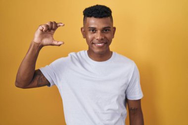 Young hispanic man standing over yellow background smiling and confident gesturing with hand doing small size sign with fingers looking and the camera. measure concept. 