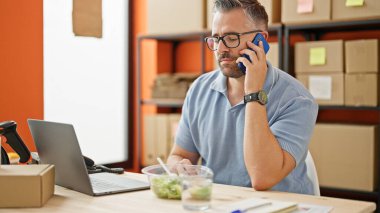 Gri saçlı adam ekomence işi. Akıllı telefondan konuşuyor. Ofiste masada oturuyor.