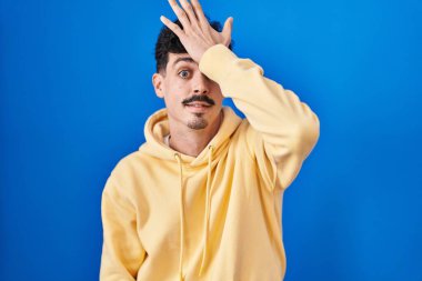 Hispanic man standing over blue background surprised with hand on head for mistake, remember error. forgot, bad memory concept. 