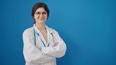 Genç, güzel İspanyol kadın doktor, gülümsüyor. Kollarını kavuşturup, mavi arka planda duruyor.
