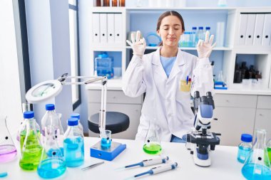 Laboratuvarda çalışan esmer bir kadın. Gözleri kapalı rahatlayıp gülümsüyor. Parmaklarıyla meditasyon yapıyor. yoga konsepti. 