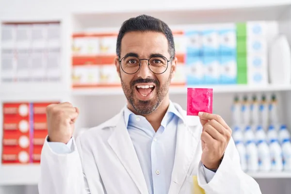 Hombre Hispano Con Barba Trabajando Farmacia Sosteniendo Preservativo Gritando Orgulloso —  Fotos de Stock