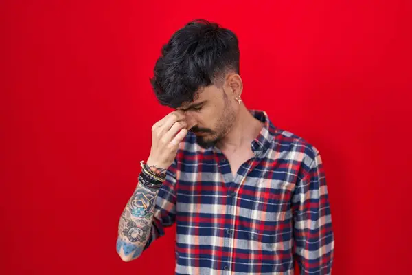 赤いバックグラウンドの上に立っているひげを持つ若いヒスパニックの男は疲労と頭痛を感じる鼻と目を疲れさせました ストレスとフラストレーションの概念 — ストック写真