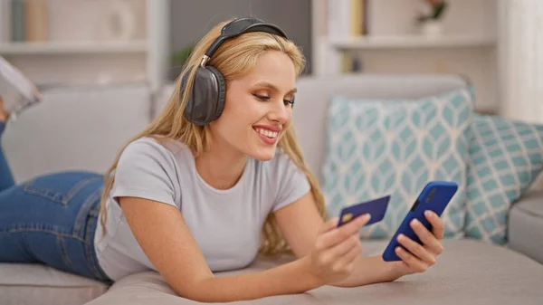 Young blonde woman shopping with smartphone and credit card listing to music at home