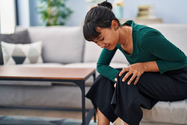 Молодая красивая латиноамериканка, сидящая дома на диване и страдающая от травмы колена