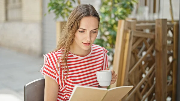Young blonde woman drinking cup of coffee reading notebook at coffee shop terrace