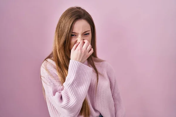 年轻的高加索女人站在粉红的背景上 闻到一股难闻 无法忍受的气味 用手指捂住鼻子呼吸 — 图库照片