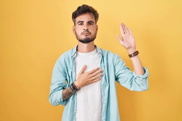 Young Hispanic Man Tattoos Standing Yellow Background Swearing Hand Chest — Stock Fotó