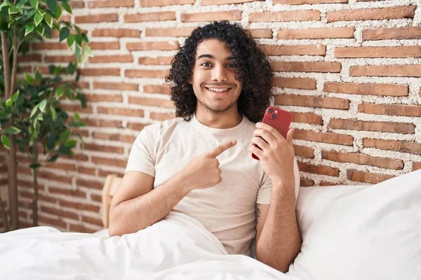 Kıvırcık Saçlı Spanyol Adam Akıllı Telefon Kullanıyor Yatakta Oturmuş Gülümsüyor — Stok fotoğraf