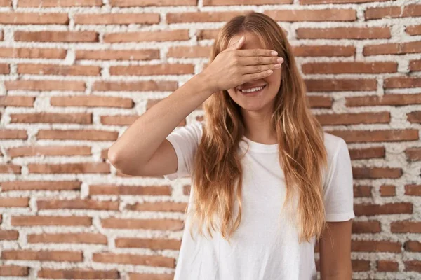 レンガの壁の上に立っている若い白人女性は笑顔で 驚きのために目を覆う顔を手で笑っています 視覚障害の概念 — ストック写真