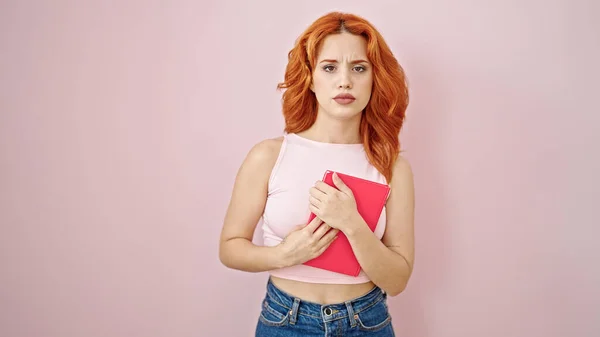 stock image Young redhead woman holding book with serious expression over isolated pink background