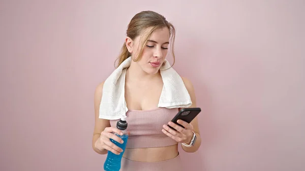 若いですブロンド女性身に着けていますスポーツウェア保持精力的な飲料使用してスマートフォン上の隔離されたピンクの背景 — ストック写真
