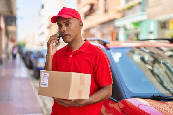 Young latin man delivery worker holding package talking on smartphone at street
