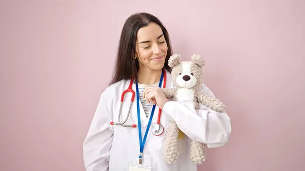 年轻美丽的惊慌失措的女医生微笑着 自信地将泰迪熊抱在孤立的粉色背景之上 — 图库照片