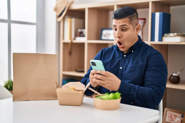Joven Hispano Comiendo Comida Para Llevar Usando Smartphone Asustado Sorprendido —  Fotos de Stock
