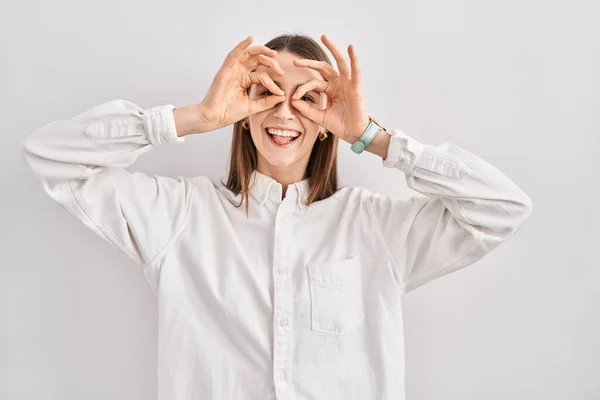 stock image Young caucasian woman standing over isolated background doing ok gesture like binoculars sticking tongue out, eyes looking through fingers. crazy expression. 