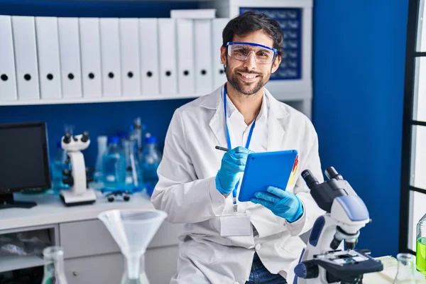 Young hispanic man scientist smiling confident using touchpad at laboratory