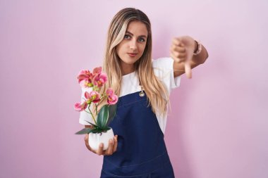 Young blonde woman wearing gardener apron holding plant looking unhappy and angry showing rejection and negative with thumbs down gesture. bad expression. 