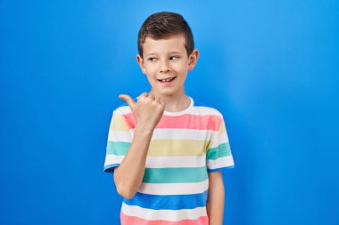 Mavi arka planda dikilen, gülümseyen, gülümseyen ve baş parmağıyla yan tarafı gösteren beyaz bir çocuk.. 