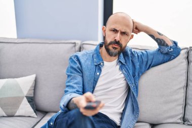 Genç kel adam televizyon seyrediyor. Evdeki sıkıcı ifadesiyle kanepede oturuyor.