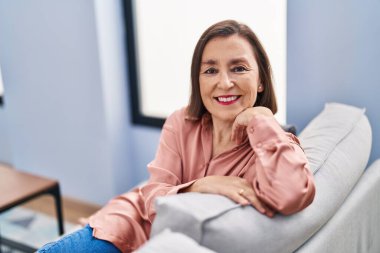 Orta yaşlı bir kadın evdeki koltukta otururken kendinden emin bir şekilde gülümsüyor.